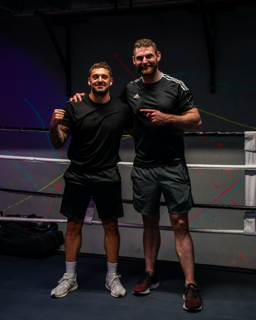 Two male boxers smiling and posing in a boxing ring, ready for training.